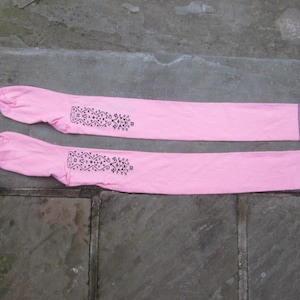 May include: Two pink arm sleeves with a decorative black floral pattern near the top. The sleeves are long and tubular, with a rounded end and a finished edge at the wrist. The sleeves are laying on a gray surface.