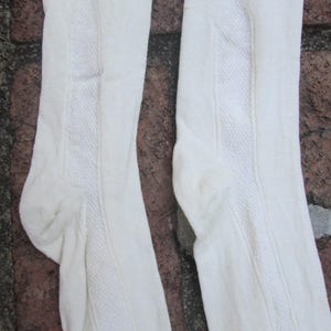 May include: Close-up of two white socks. The socks are made of a soft material and have a textured pattern along the sides and down the front. The socks are slightly off-white in color and appear to be new.
