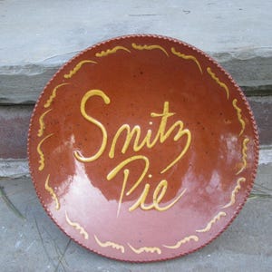 May include: A round, reddish-brown ceramic plate with the words "Snitz Pie" in yellow script. The plate has a decorative wavy border and is sitting on a stone surface.