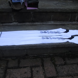 May include: Two white over-the-knee socks with black accents and floral embroidery. Each sock has a black band at the top and three black stripes near the top. The socks are laid out on a stone surface.