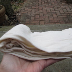 Op de afbeelding: Een gevouwen witte katoenen stof, mogelijk een theedoek of een vaatdoek, wordt in een hand gehouden.