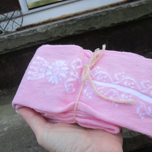 May include: A stack of pink socks tied with a light brown string. Each sock has a white floral design and a white stripe. The socks are held in a hand.