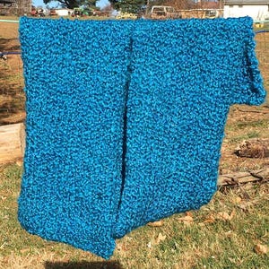 Op de afbeelding: Twee turquoise blauwe gebreide sjaals hangen buiten aan een waslijn. De sjaals zijn gemaakt van een pluche, getextureerd garen.