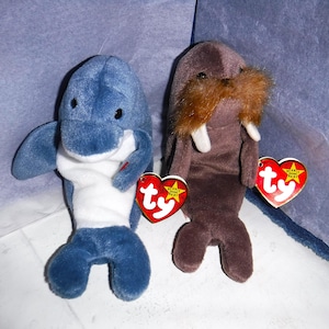 May include: Two plush toys, a blue dolphin and a brown walrus, with red heart-shaped Ty tags attached. The dolphin has a white belly and a toothy grin. The walrus has a brown mustache and white tusks.