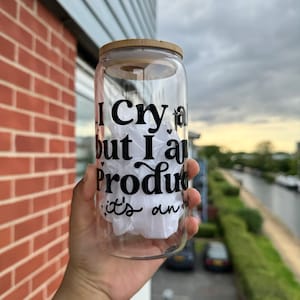 May include: A clear glass can-shaped tumbler with a bamboo lid. The tumbler features black text: "I cry a lot but I am productive it's an". The glass is filled with white paper.