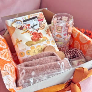May include: A gift box containing a glass tumbler with a cosy books print, a fuzzy mauve headband, a brown and white checkered scrunchie, a small candle, and a wrapped item with a wax seal, all nestled in pumpkin-patterned tissue paper.