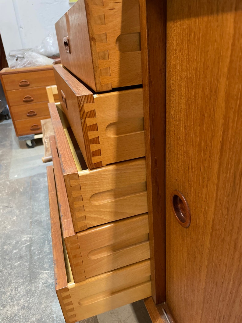 May include: A wooden dresser with five drawers and a sliding door. The drawers are open and show the dovetail joinery. The dresser is made of a light wood with a natural finish.