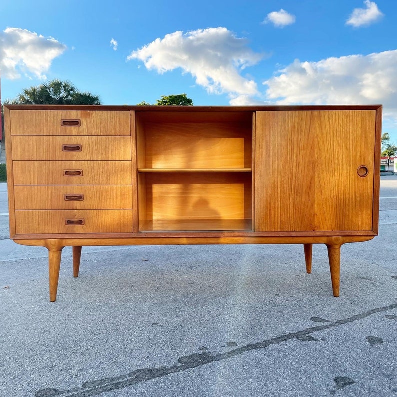 May include: A mid-century modern teak sideboard with four drawers and a cabinet with a shelf. The cabinet has a sliding door. The sideboard has tapered legs.