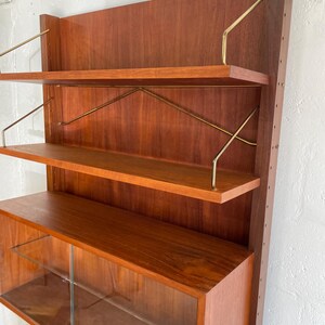 Op de afbeelding: Een modern wandmeubel in mid-century stijl, gemaakt van hout met glazen planken en messing accenten. Het meubel heeft drie open planken en een onderkast met glazen deuren.