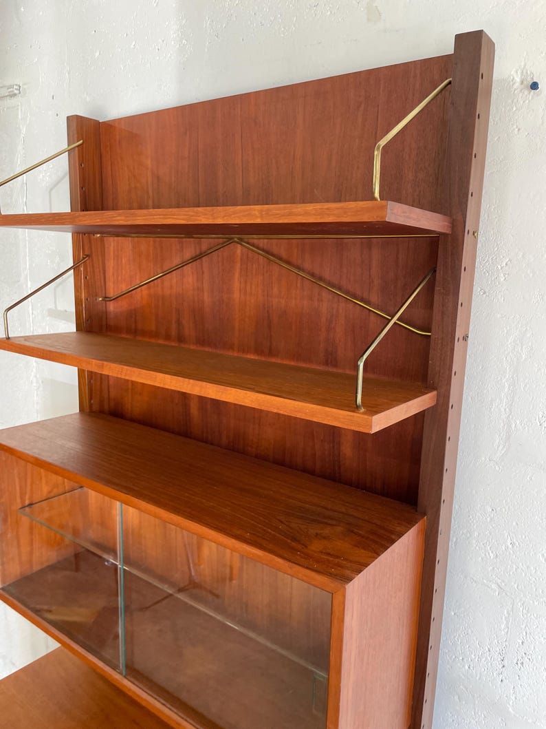 Op de afbeelding: Een modern wandmeubel in mid-century stijl, gemaakt van hout met glazen planken en messing accenten. Het meubel heeft drie open planken en een onderkast met glazen deuren.