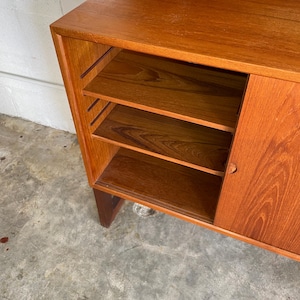 Op de afbeelding: Een houten kast met een schuifdeur en open planken. De kast is gemaakt van teakhout en heeft een natuurlijke afwerking. De planken zijn verstelbaar en kunnen worden gebruikt om boeken, serviesgoed of andere items op te bergen.