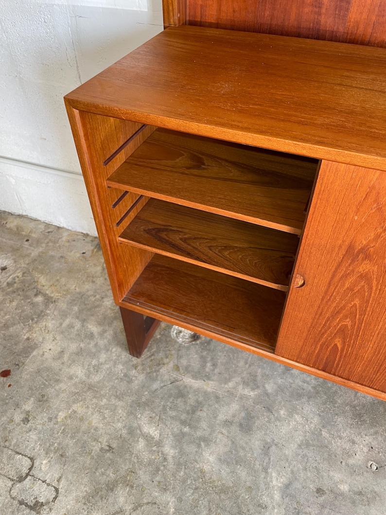 Op de afbeelding: Een houten kast met een schuifdeur en open planken. De kast is gemaakt van teakhout en heeft een natuurlijke afwerking. De planken zijn verstelbaar en kunnen worden gebruikt om boeken, serviesgoed of andere items op te bergen.