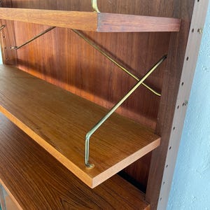 Op de afbeelding: Een houten wandunit met planken en een kast. De planken worden ondersteund door messing staven. De kast heeft een glazen deur.
