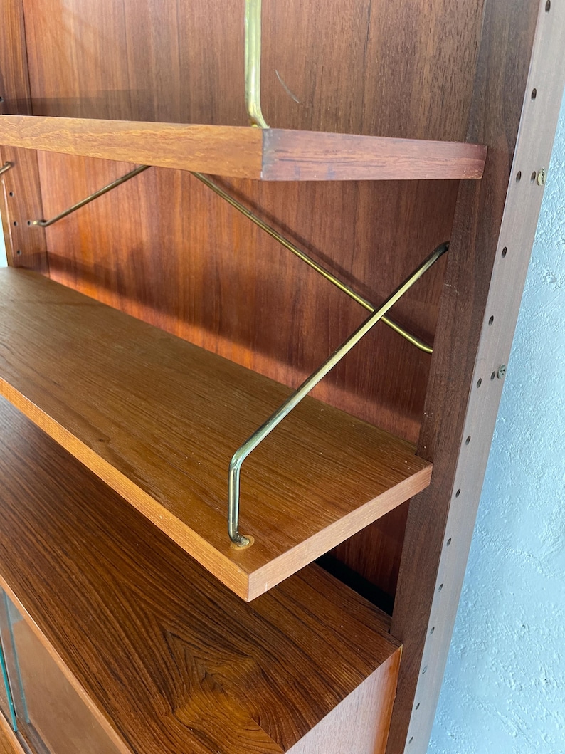 Op de afbeelding: Een houten wandunit met planken en een kast. De planken worden ondersteund door messing staven. De kast heeft een glazen deur.