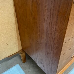 May include: A close-up of the corner of a wooden cabinet with drawers. The cabinet is made of a light brown wood with a natural grain pattern. The drawers are made of the same wood and have a simple design.