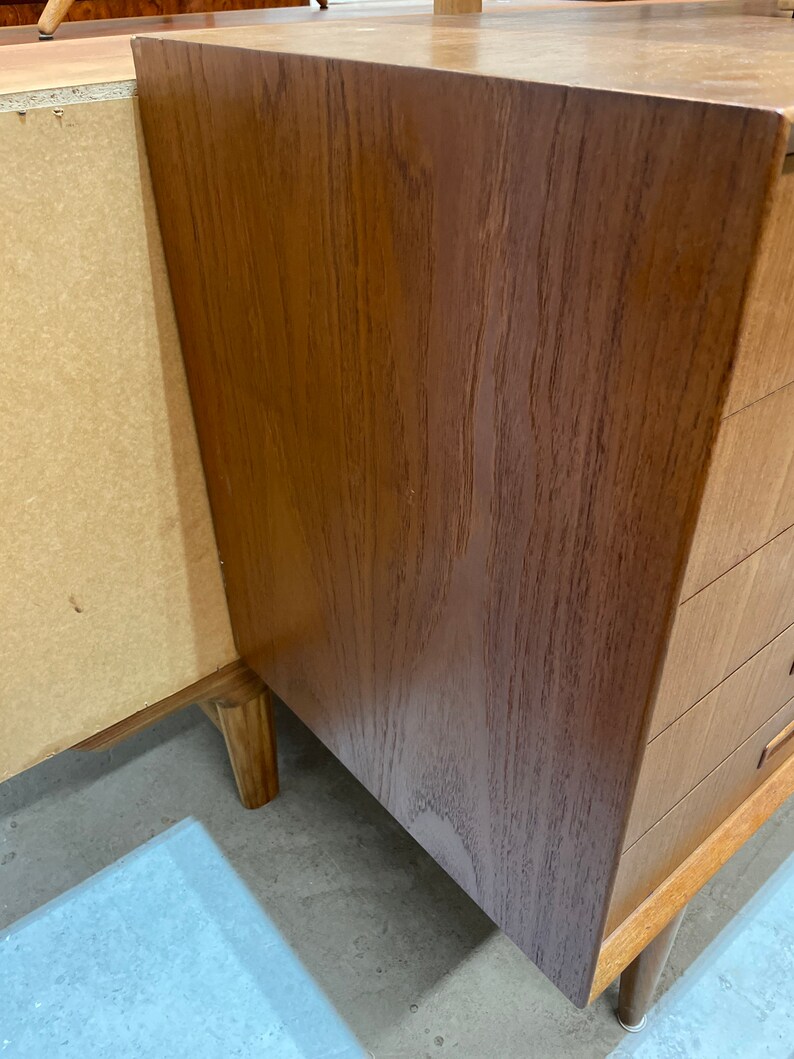 May include: A close-up of the corner of a wooden cabinet with drawers. The cabinet is made of a light brown wood with a natural grain pattern. The drawers are made of the same wood and have a simple design.
