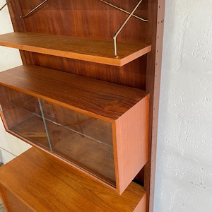 Op de afbeelding: Een moderne wandunit van teak uit het midden van de eeuw met glazen deuren en planken. De unit heeft een kast met een lade aan de onderkant.