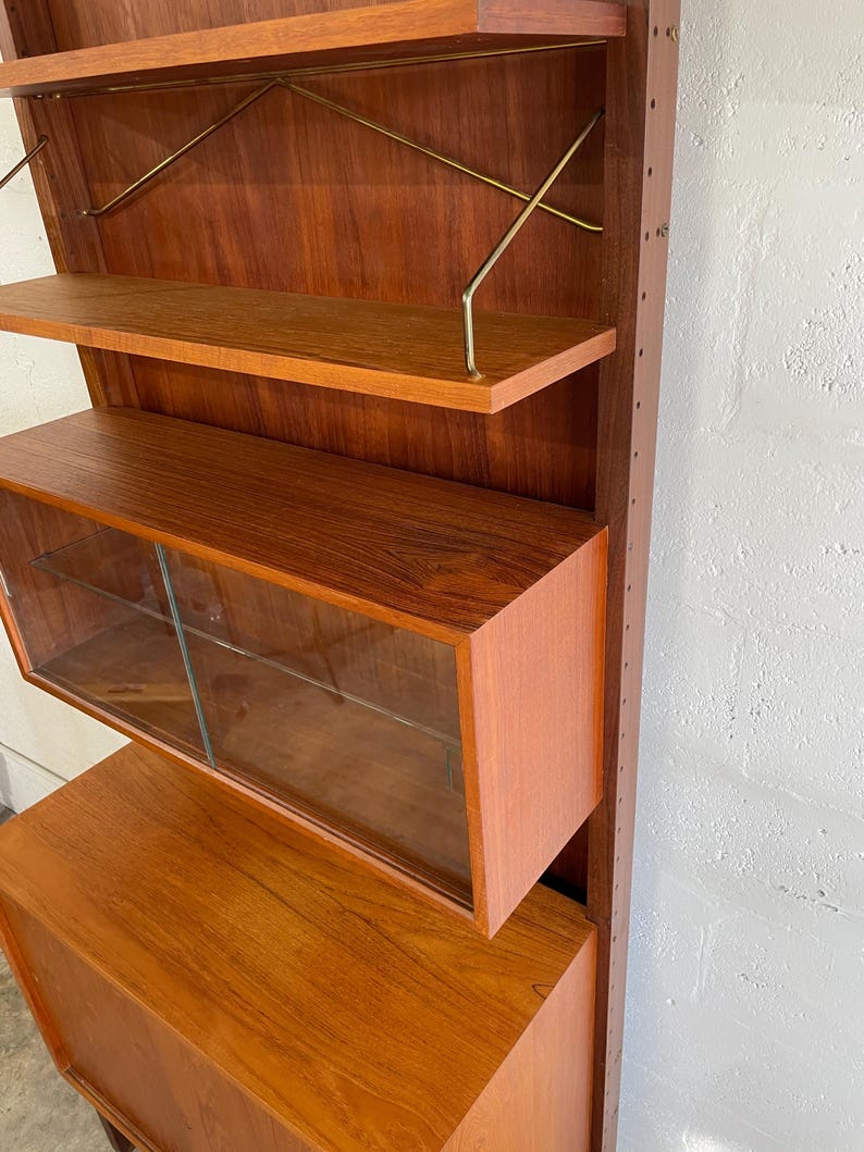 Op de afbeelding: Een moderne wandunit van teak uit het midden van de eeuw met glazen deuren en planken. De unit heeft een kast met een lade aan de onderkant.