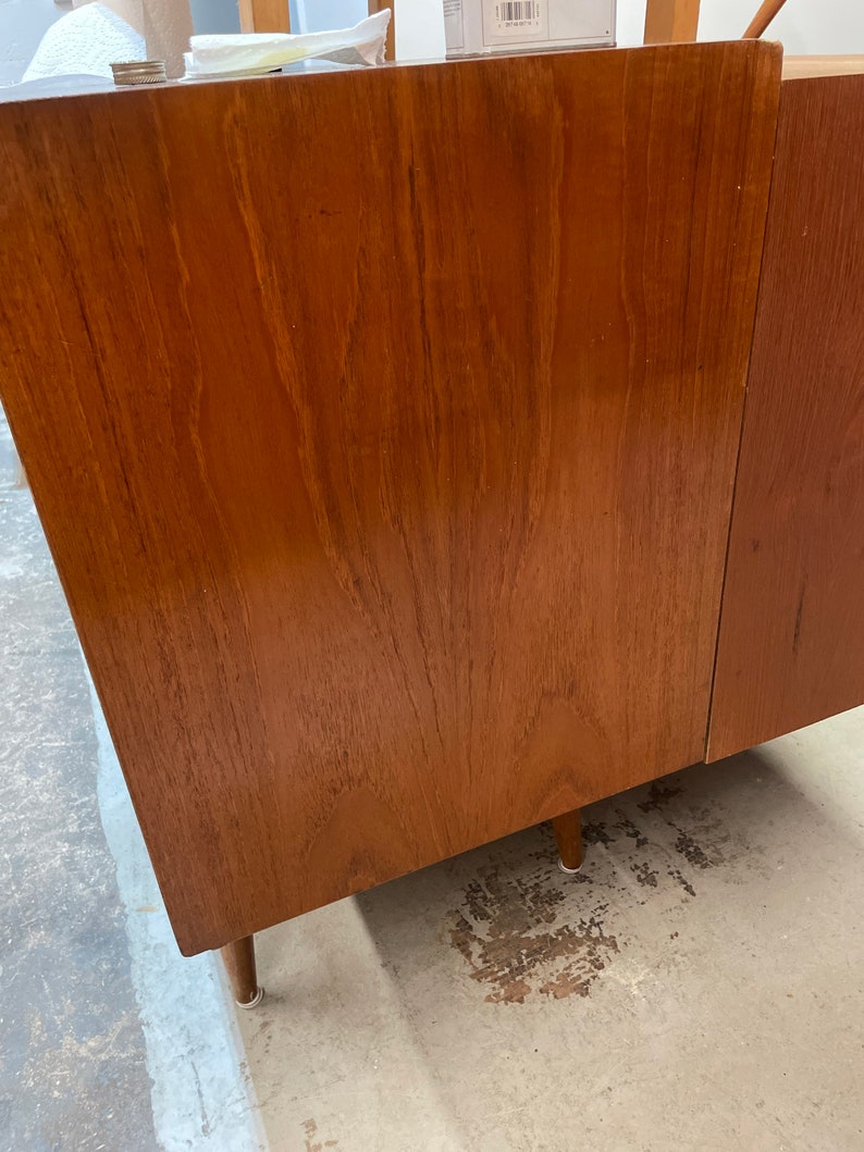 May include: A close-up of a wooden cabinet with a light brown finish. The cabinet has a simple design with a smooth surface and visible wood grain. The cabinet is sitting on a light gray floor.
