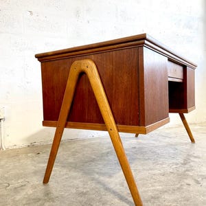 May include: Mid-century modern wooden desk with a warm brown finish. The desk features a rectangular top with a drawer and a unique A-frame leg design. The legs are made of a lighter wood, creating a contrast with the desk's body.