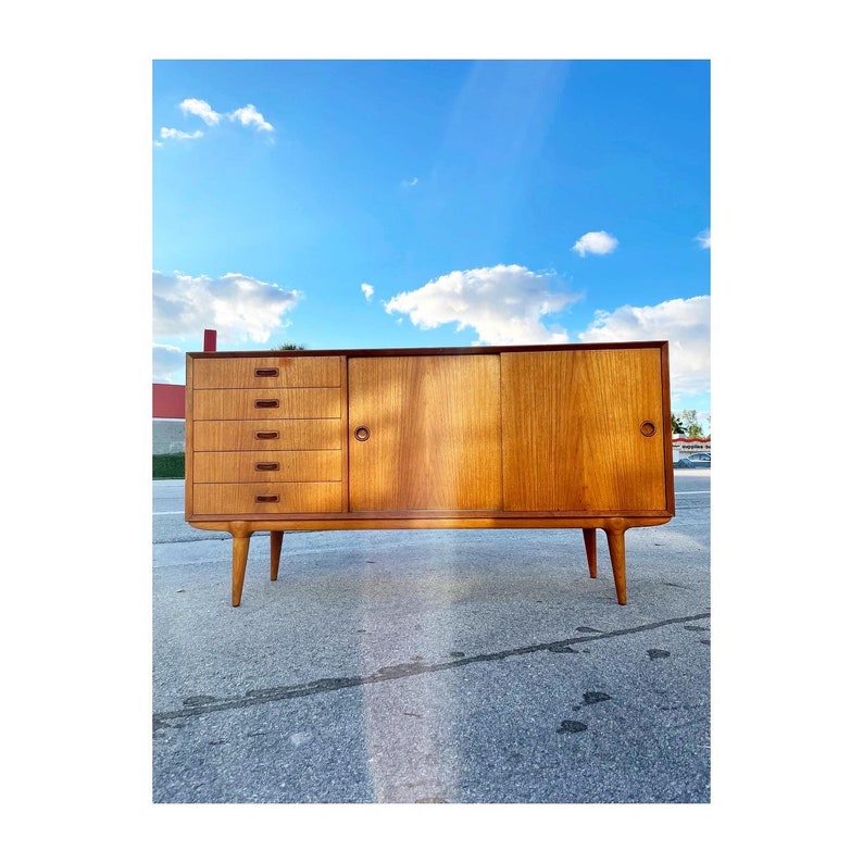 May include: A mid-century modern teak sideboard with four drawers and two doors. The sideboard has a light wood finish and sits on four tapered legs.