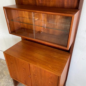Op de afbeelding: Een hoge houten kast met glazen deuren en planken. De kast heeft een lager gedeelte met twee massieve deuren. De kast is gemaakt van een rijk, roodbruin hout.