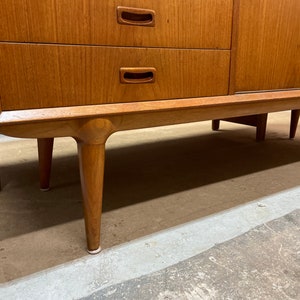 May include: A vintage wooden sideboard with four drawers and two doors. The sideboard has a light wood finish and features simple, rectangular handles. The sideboard is sitting on four tapered legs.