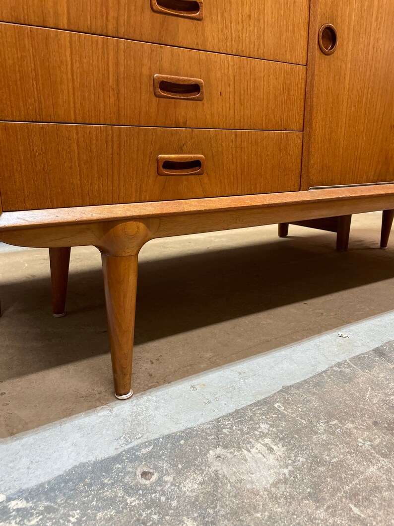 May include: A vintage wooden sideboard with four drawers and two doors. The sideboard has a light wood finish and features simple, rectangular handles. The sideboard is sitting on four tapered legs.