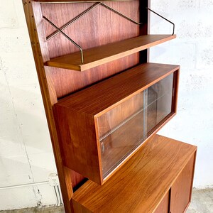 Op de afbeelding: Een moderne wandunit van teak uit het midden van de eeuw met een vitrinekast, twee planken met messing steunen en een onderkast met twee deuren. De unit is gemaakt van massief teak en heeft een warme, natuurlijke afwerking.