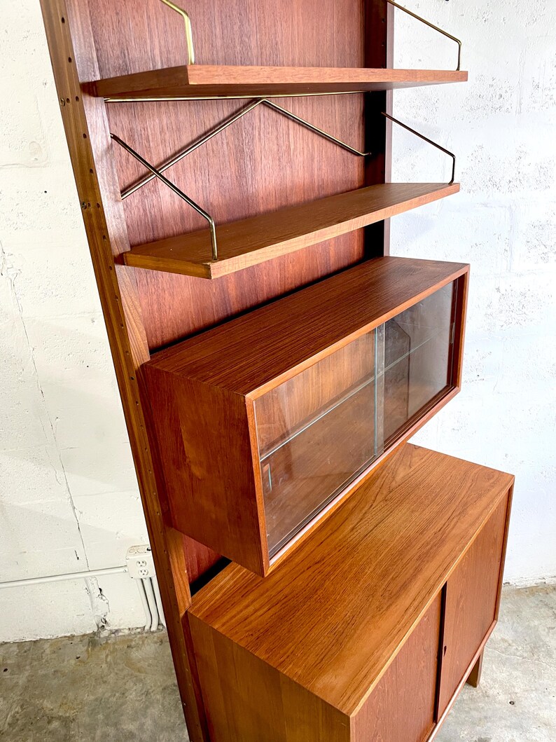 Op de afbeelding: Een moderne wandunit van teak uit het midden van de eeuw met een vitrinekast, twee planken met messing steunen en een onderkast met twee deuren. De unit is gemaakt van massief teak en heeft een warme, natuurlijke afwerking.