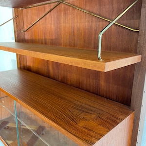 Op de afbeelding: Een mid-century modern teak wandunit met glazen deuren en planken. De unit heeft een warme bruine houtkleur en messing accenten.