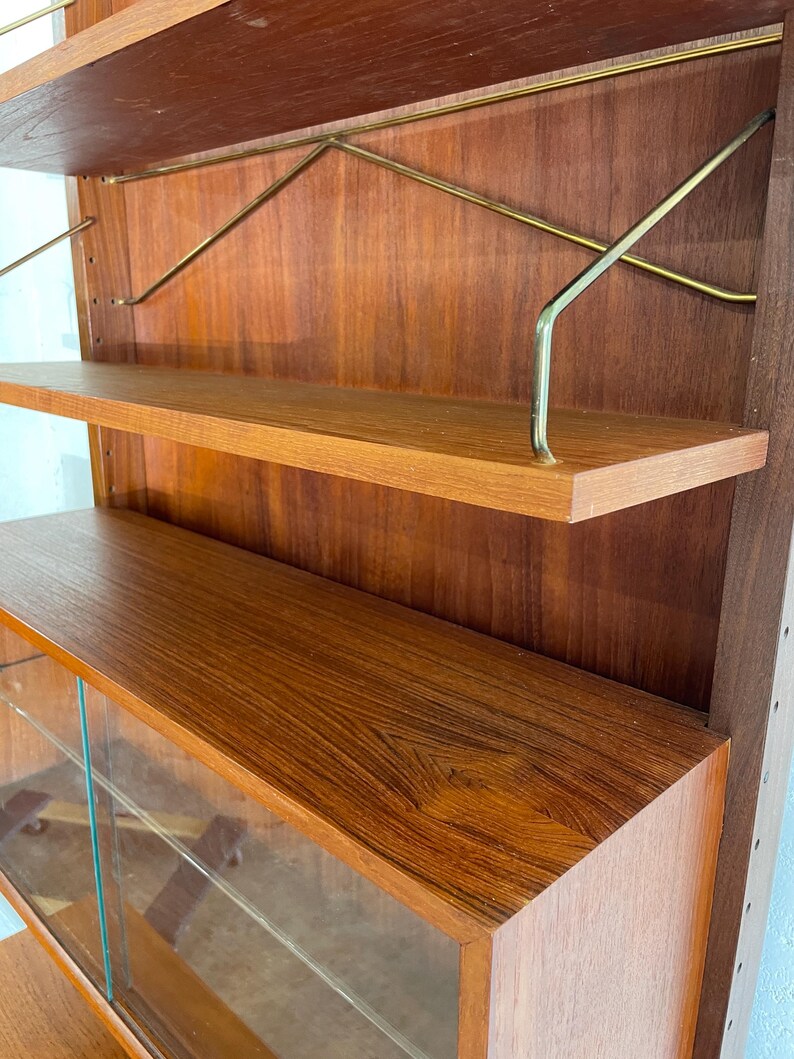 Op de afbeelding: Een mid-century modern teak wandunit met glazen deuren en planken. De unit heeft een warme bruine houtkleur en messing accenten.