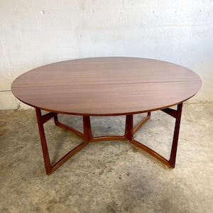 May include: A round, mid-century modern wooden table with a warm brown finish. The table has a split top and a unique base with angled legs. The table is approximately 48 inches in diameter.