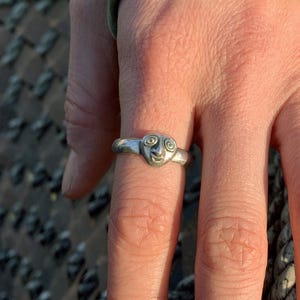 May include: A silver ring with a spiral design is worn on a finger. The ring is the focal point, set against a backdrop of a person's hand, a green sleeve, and a woven rope. The ring's design is detailed and eye-catching.
