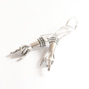 May include: Silver-toned earrings featuring hand gestures, with the index and pinky fingers extended, forming a rock and roll symbol. The earrings have a detailed design, with a hook closure. The jewelry is set against a white background.