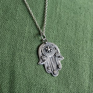 May include: A silver-colored Hamsa hand pendant necklace. The pendant features intricate detailing and hangs from a delicate chain. The background is a textured green fabric.