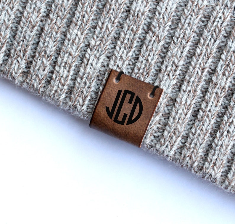 May include: A brown leather label with a black circle monogrammed with the letters "JCD" attached to a gray knitted beanie.