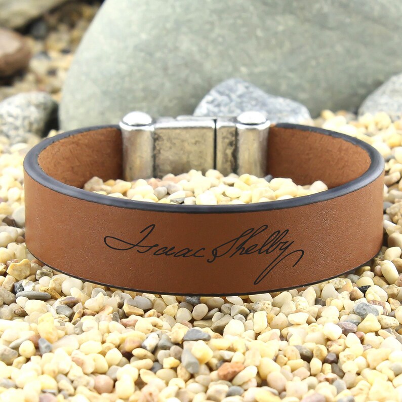 May include: A brown leather bracelet with a silver magnetic clasp. The bracelet is engraved with the name "Isaac Shelly" in cursive.