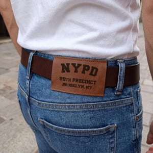 May include: A brown leather belt with a rectangular patch displaying "NYPD 99th Precinct Brooklyn, NY" is worn with blue denim jeans. The belt is fastened with a silver buckle. The person is wearing a white t-shirt.