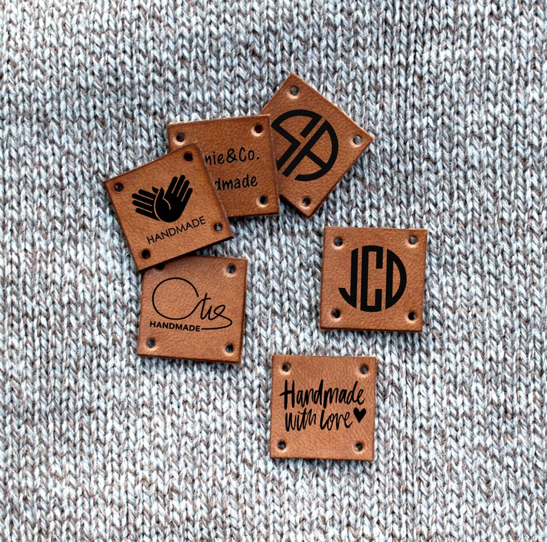 May include: A collection of square, brown leather tags with black text and designs. The tags feature phrases like "Handmade" and "Handmade with love," along with initials and logos. Each tag has four corner holes for sewing.