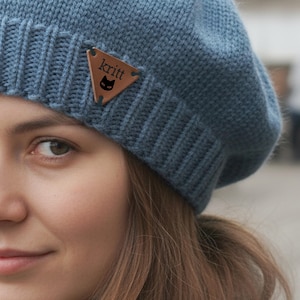 May include: A blue knit beret with a brown leather triangle tag. The tag features the word "kritt" and a black cat silhouette. The beret is made of a soft, textured material.