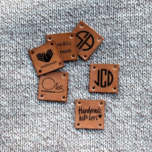 May include: Five brown leather labels with different designs. The labels have holes punched in the corners and are on a gray knitted fabric background. The labels say "Handmade", "Handmade with Love", "J.C.D.", "Otis Handmade", and "nie & Co. Handmade".