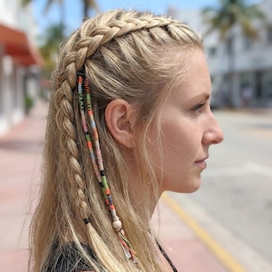 Op de afbeelding: Blond haar gestyled in een zijvlecht met een kroonvlecht. Kleurrijke, patroon haar wraps zijn in de zijvlecht geweven. De achtergrond is wazig en toont een straatbeeld met palmbomen.