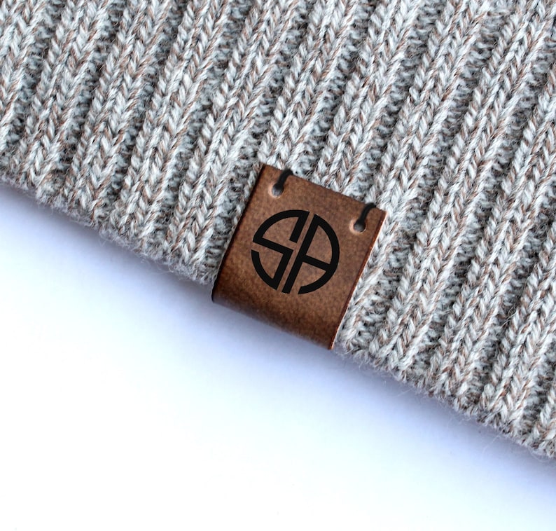 May include: A brown leather label with a black circle containing the letters "SB" in a sans-serif font. The label is attached to a gray knitted fabric.