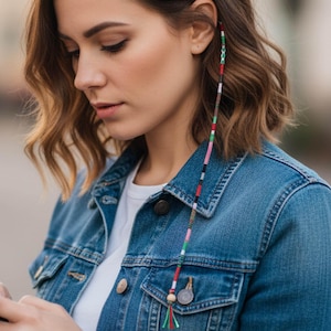 May include: A colorful hair wrap with a beaded design, featuring red, green, white, and black accents. The hair accessory is worn near the ear and hangs down. The person is wearing a denim jacket.