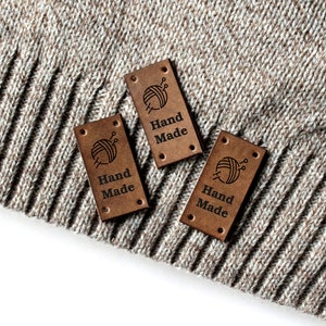May include: Three brown leather labels with the text "Hand Made" and a small graphic of a ball of yarn and knitting needles. The labels are on a beige knitted fabric.