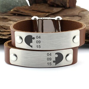 May include: Two brown leather bracelets with silver metal plates. Each plate has a half heart puzzle piece design and the numbers 04 09 15.