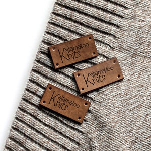 May include: Three brown leather labels with the text "Kalamazoo Knits" in black lettering. The labels are on a beige knitted fabric.