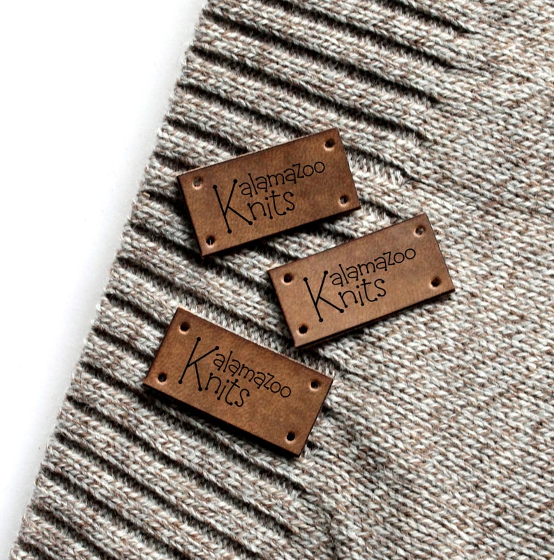 May include: Three brown leather labels with the text "Kalamazoo Knits" in black lettering. The labels are on a beige knitted fabric.
