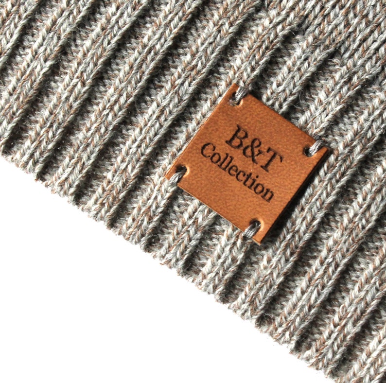 Pode incluir: Close-up de uma etiqueta de couro marrom com o texto "B&T Collection" em um tecido de malha cinza.